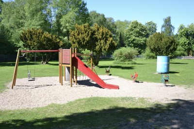 Spielplatz mit Schaukel, Klättergerüst und Rutsche mit Bäumen im Hintergrund.