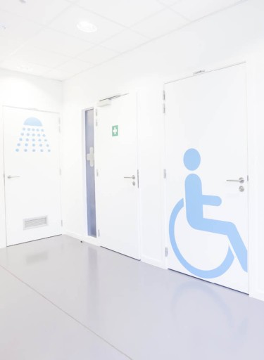 Flur mit weißen Türen und blauem Strichmännchen im Rollstuhl auf einer Tür abgebildet