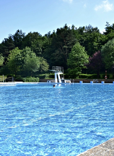 Freibadbecken mit Sprungturm in wäldlicher Umgebung.