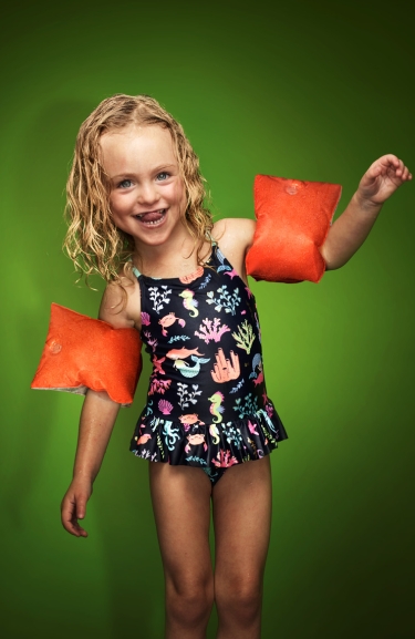 Kleines blondes, fröhliches Mädchen im Blümchenbadeanzug und Schwimmflügeln.