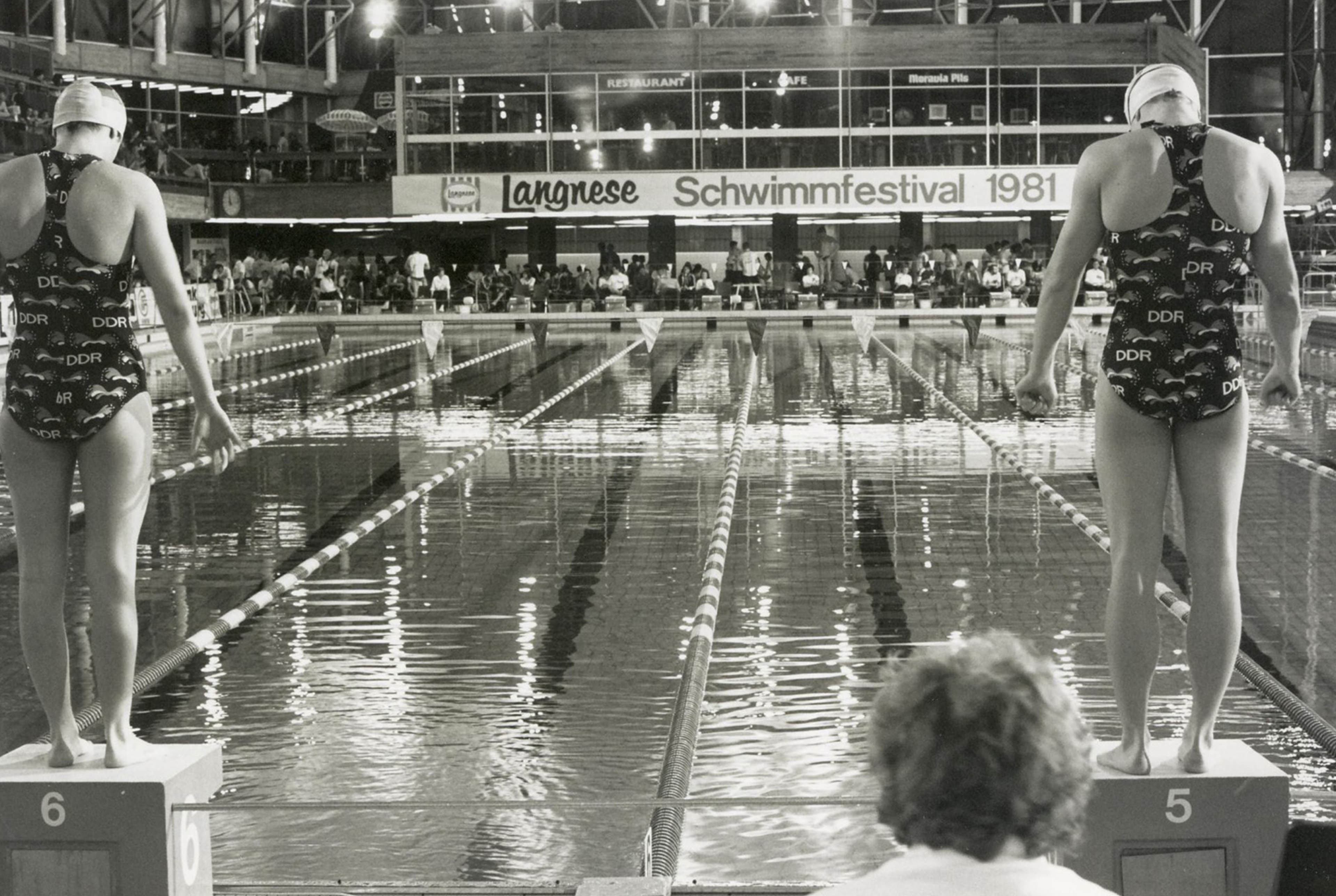 Zwei Frauen stehen in DDR-Badeanzügen auf Startblöcken vor einem Schwimmbecken