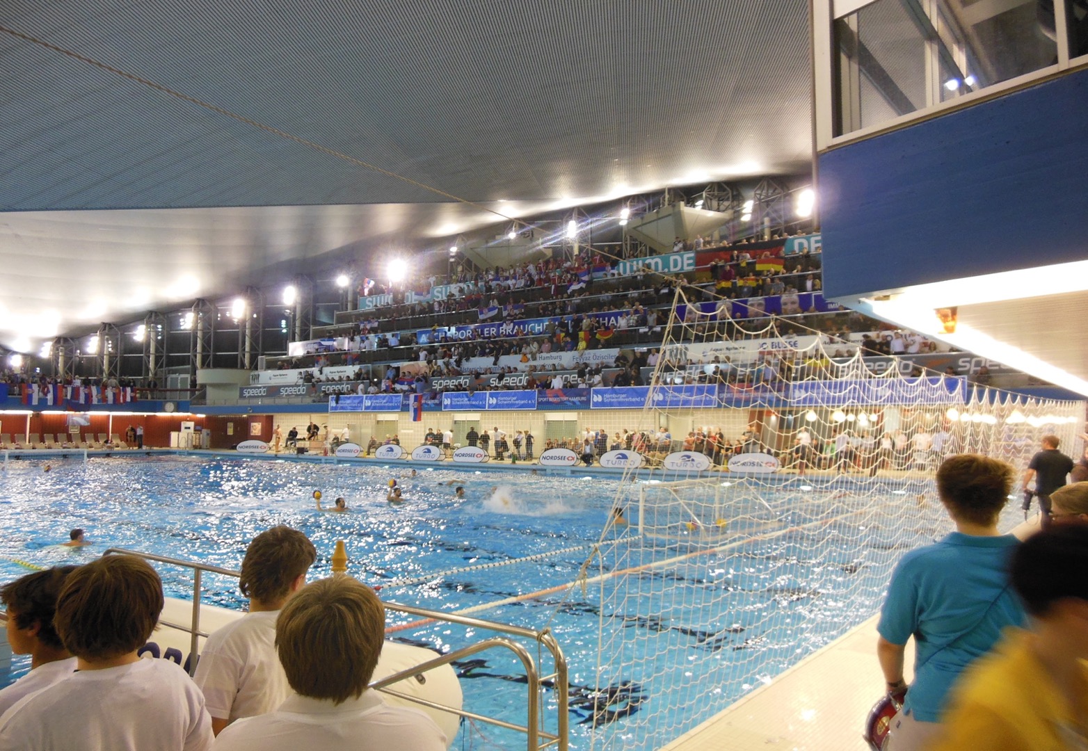 Schwimmbecken mit Männern die Bälle werfen und von Menschen auf der Tribüne beobachtet werden