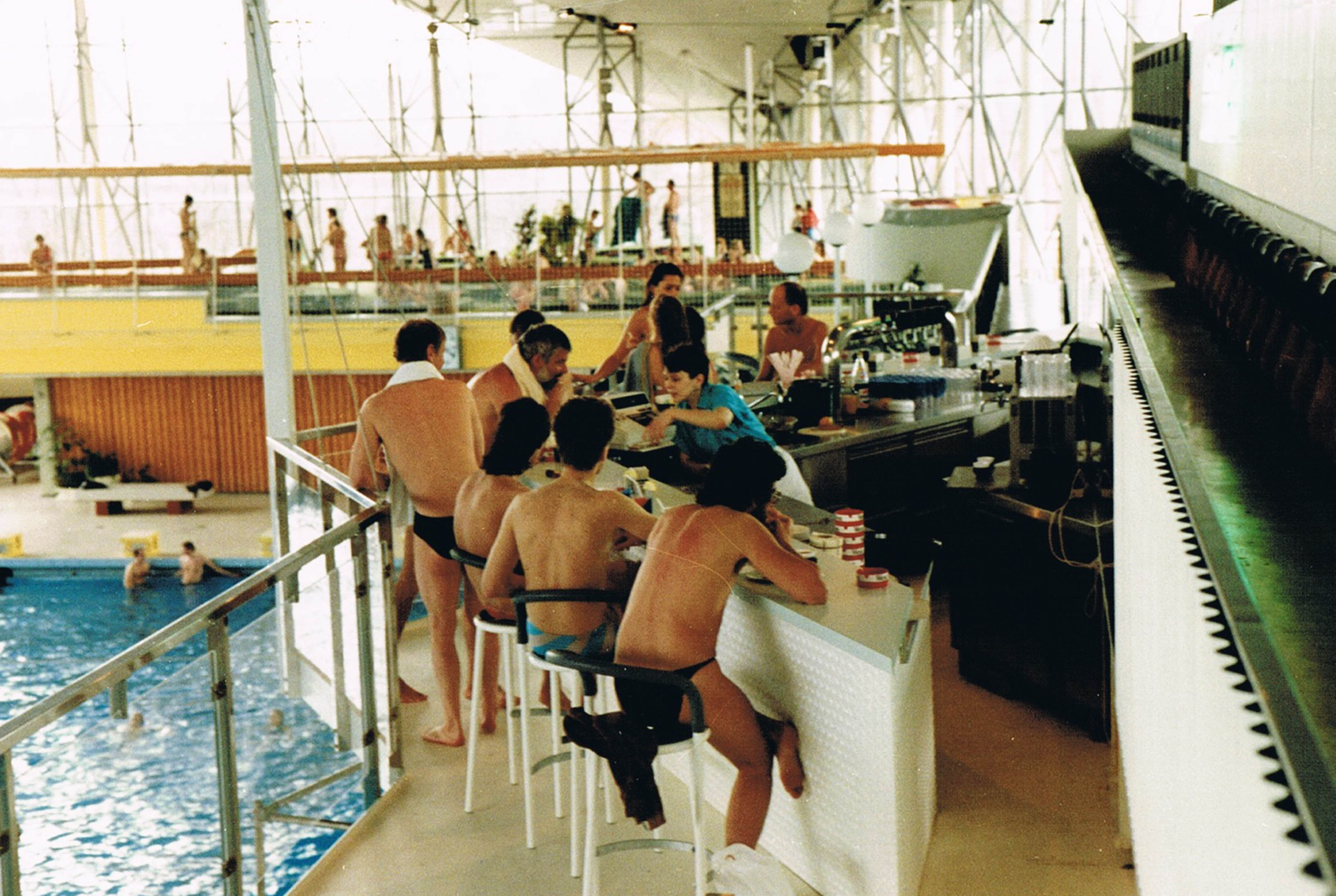 Personen stehen mit Getränken an einer Bar im Schwimmbad und blicken auf das eine Ebene tieferliegende Schwimmbecken