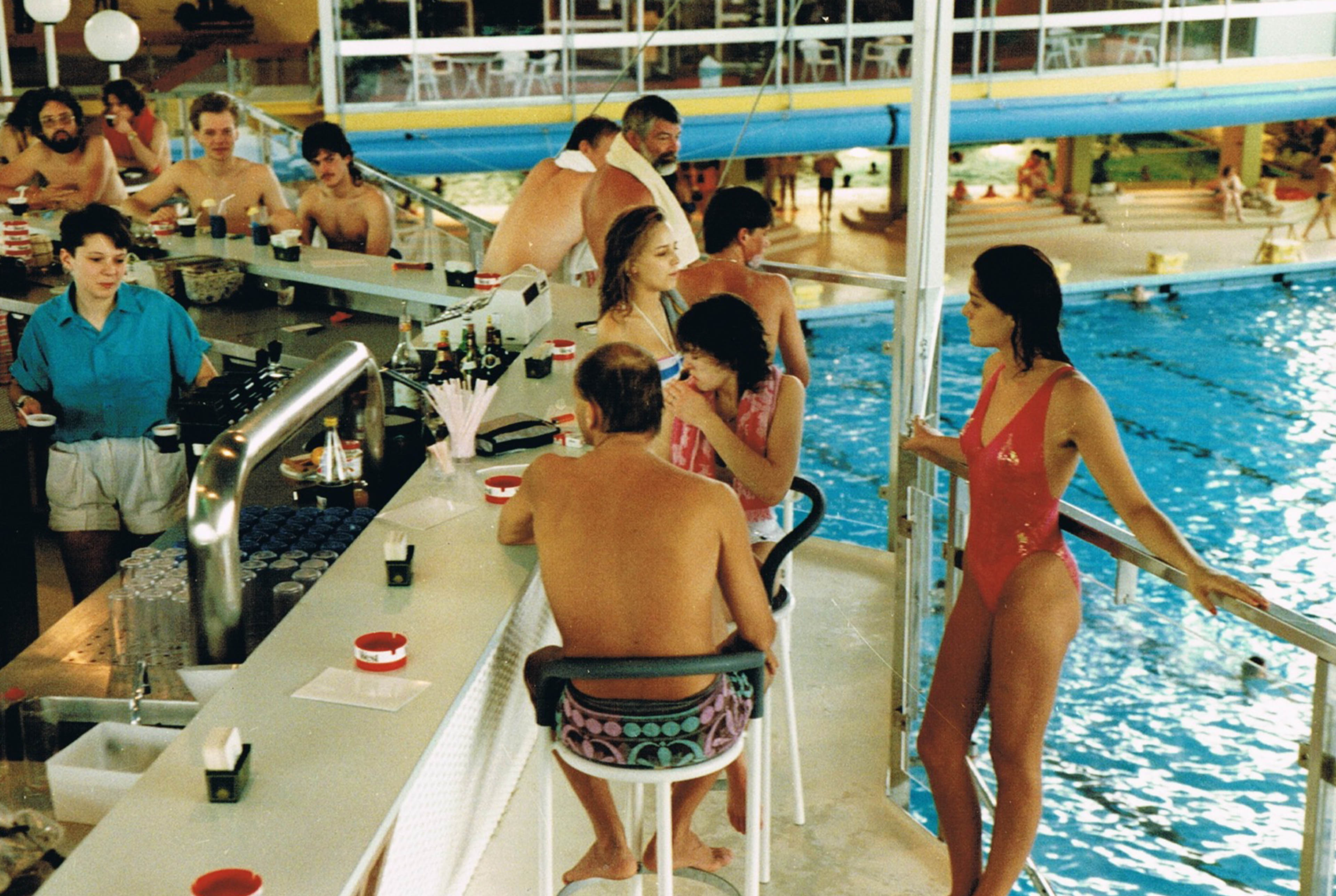 Personen stehen mit Getränken an einer Bar im Schwimmbad und blicken auf das eine Ebene tieferliegende Schwimmbecken