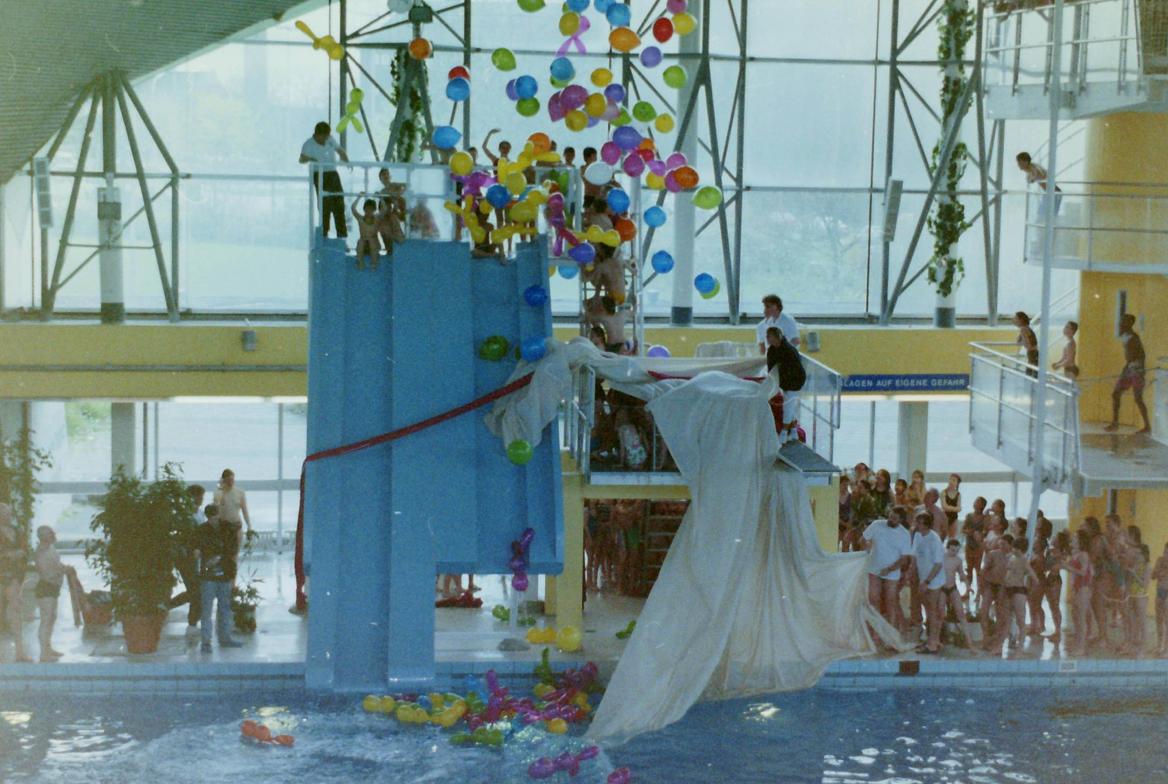 Einige Menschen stehen auf einem Sprungturm uns einer Rutsche. Luftballons fliegen umher.