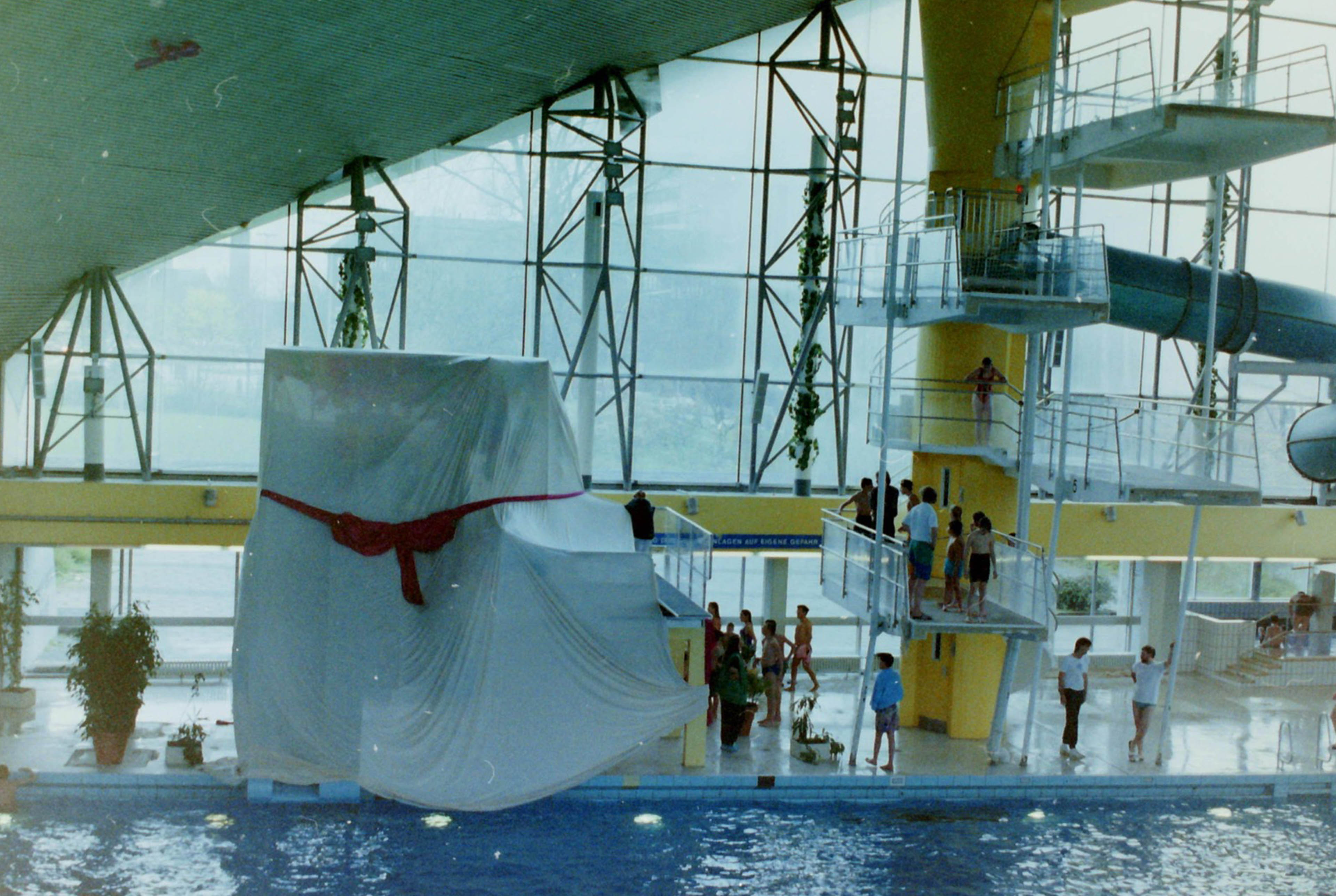 Blick auf ein Schwimmbecken mit Sprungturm. Auf und neben dem Sprungturm stehen Personen. Daneben überdeckt ein weißes Tuch mit roter Schleife etwas großes als Überraschung.