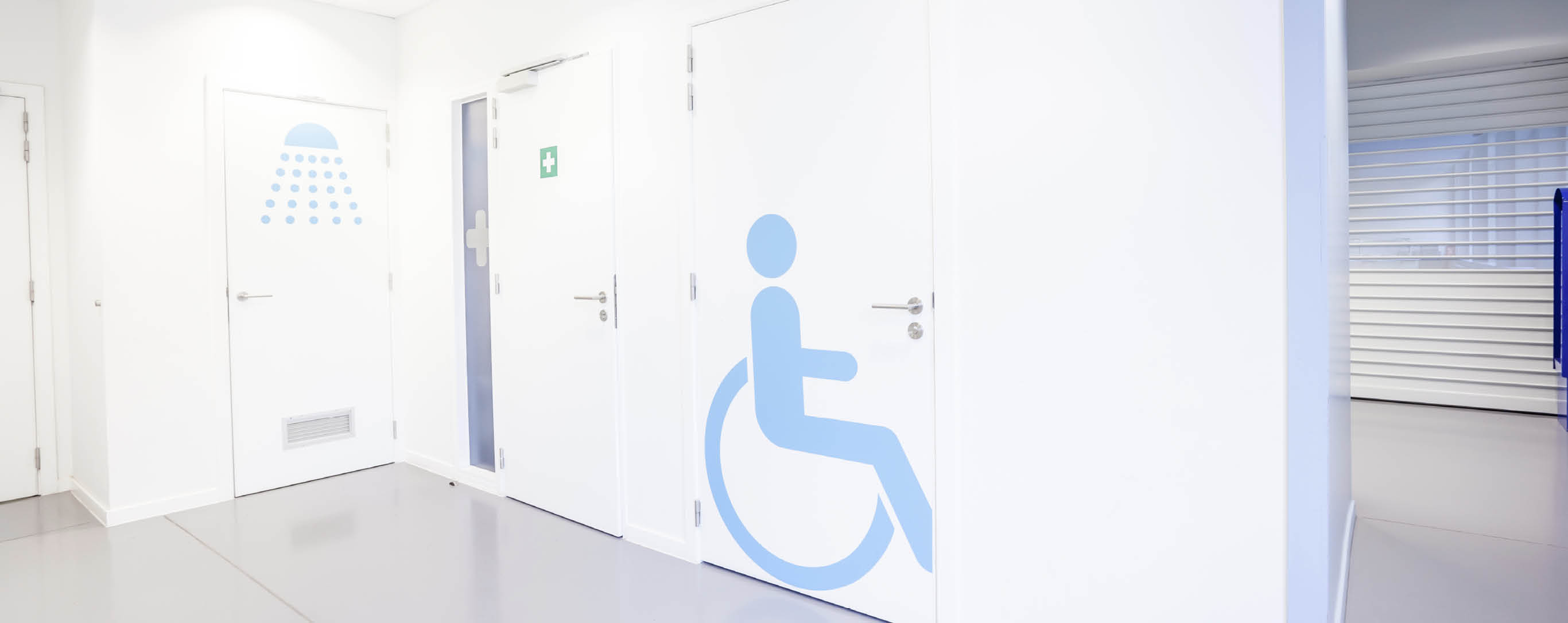 Flur mit weißen Türen und blauem Strichmännchen im Rollstuhl auf einer Tür abgebildet