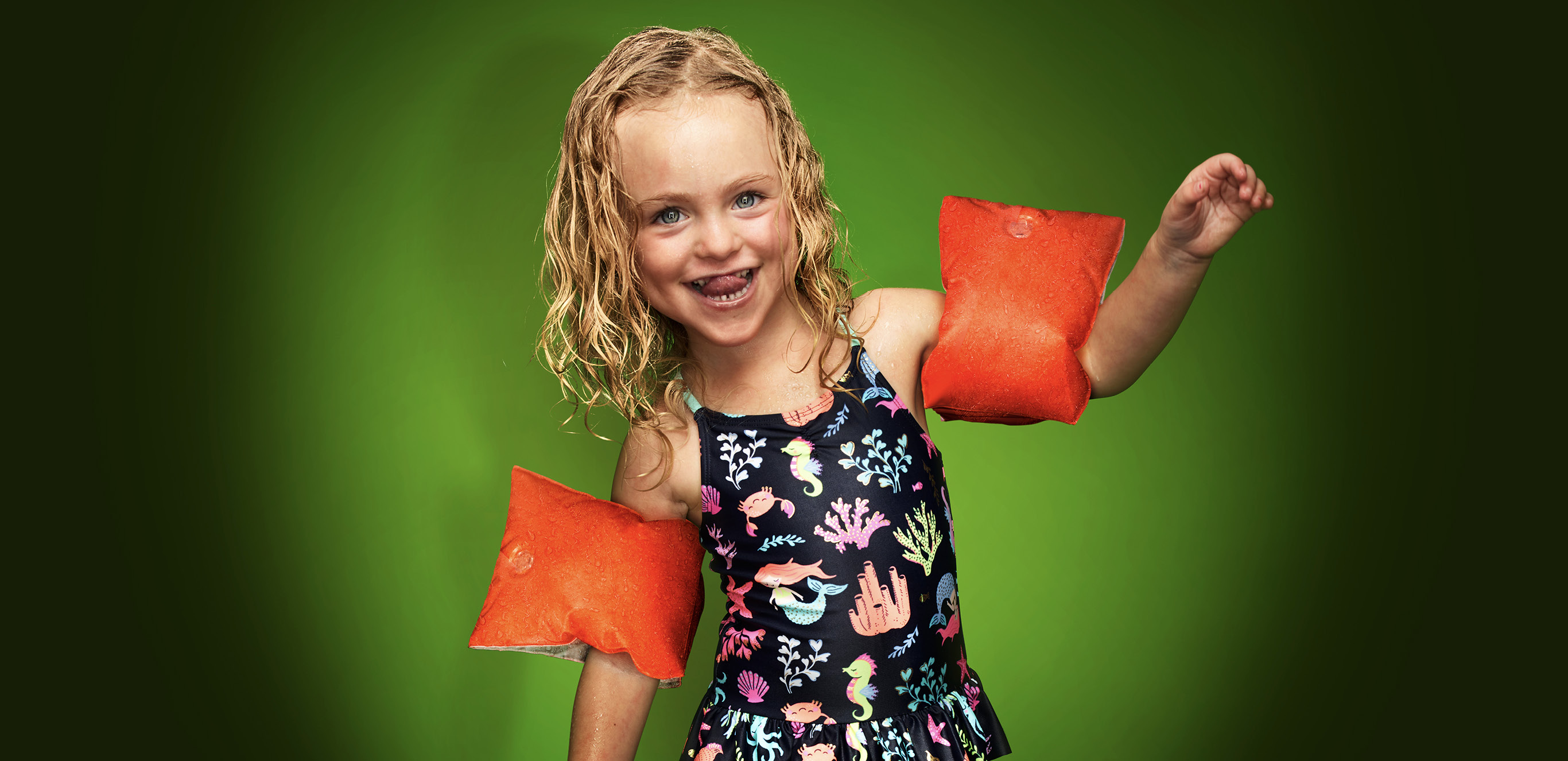 Kleines blondes, fröhliches Mädchen im Blümchenbadeanzug und Schwimmflügeln.
