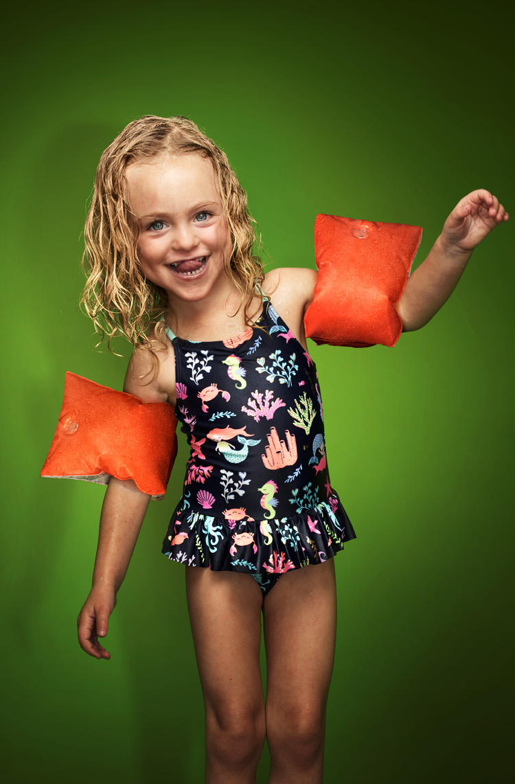 Kleines blondes, fröhliches Mädchen im Blümchenbadeanzug und Schwimmflügeln.