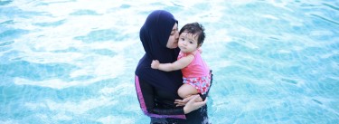 Frau im Burkini hält ein Baby auf dem Arm und steht im Wasser.