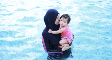 Frau im Burkini hält ein Baby auf dem Arm und steht im Wasser.