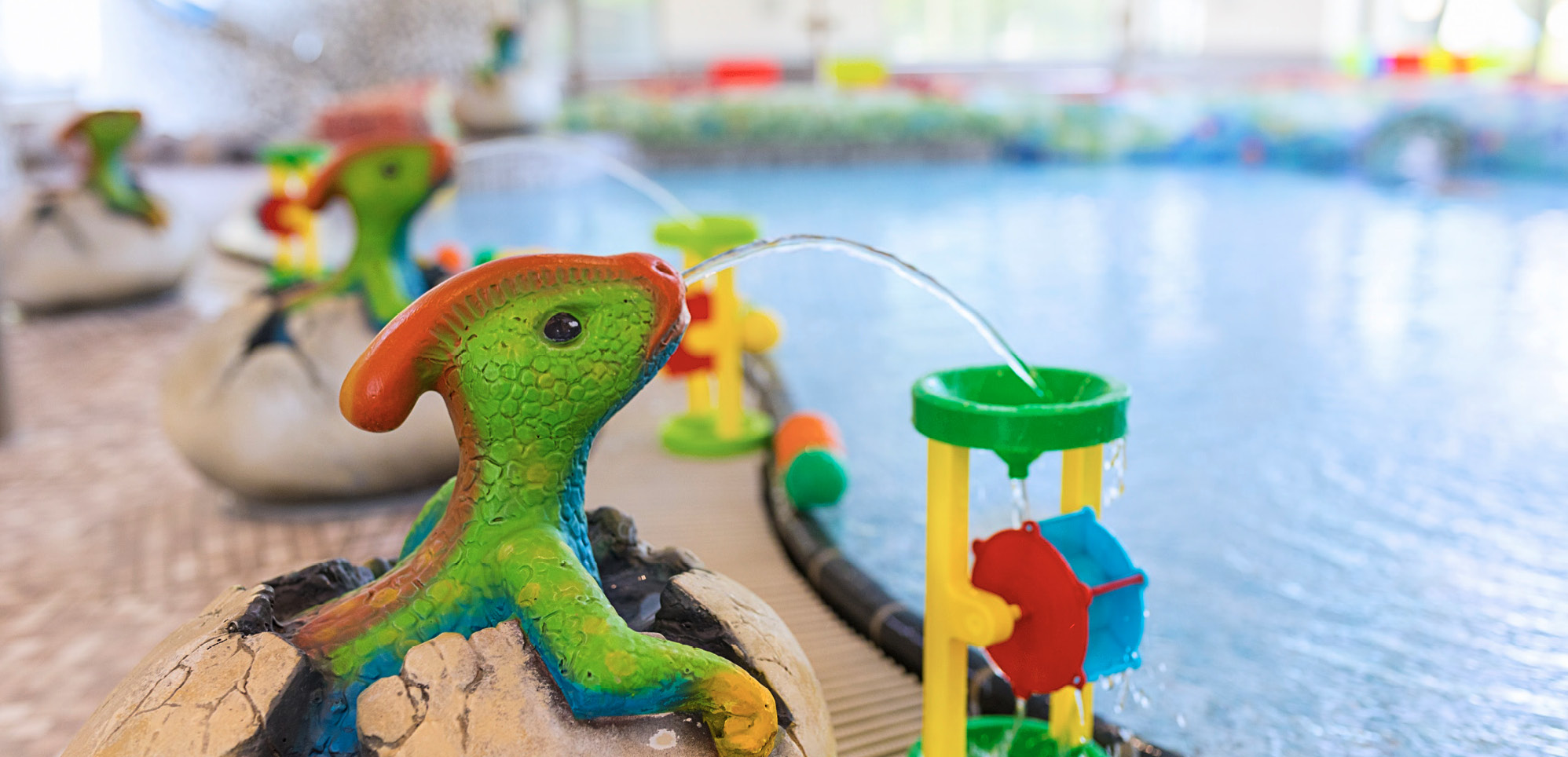 Nahaufnahme des Beckenrands aus dem Kinderbereich. Wasserspuckende Dinosaurier setzen festinstaliertes Kinderspielzeug durch Wasser in Bewegung.