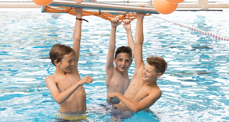 Drei Jungs hängen fröhlich an einem Klettergerüst im und über dem Wasser.