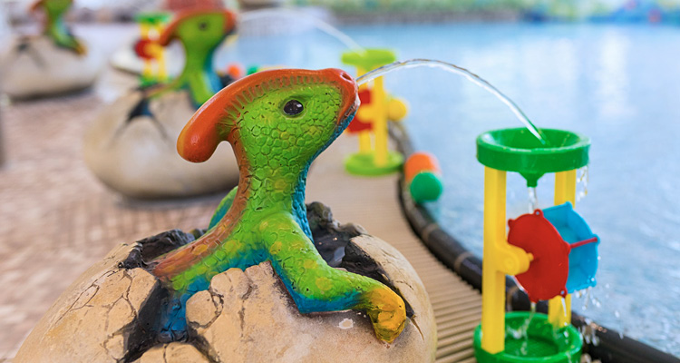 Nahaufnahme des Beckenrands aus dem Kinderbereich. Wasserspuckende Dinosaurier setzen festinstaliertes Kinderspielzeug durch Wasser in Bewegung.