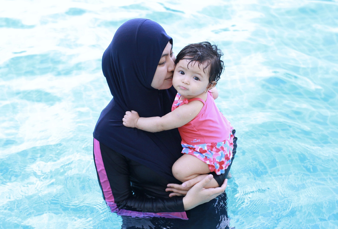 Frau im Burkini hält ein Baby auf dem Arm und steht im Wasser.