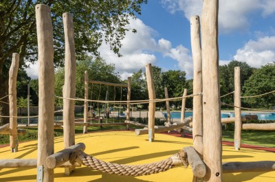 Aufnahme aus dem Außenbereich. Mehrere Holzpfähle ergeben zusamme einen Kletterbereich für Kinder. Im Hintergrund sind Schaukeln und ein Schwimmbecken zu erkennen.