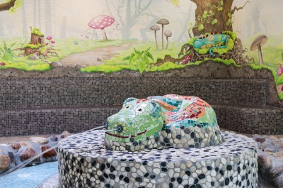 Nahaufnahme aus dem Kinderbereich. Im Hintergrund eine mit Blumen, Pilzen und Bäumen bemalte Wand. Im Vordergrund ruht ein buntes, wasserspeiendes Krokodil auf einer Erhöhung.