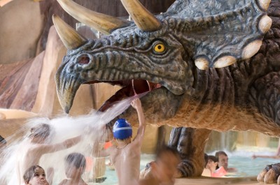 Viele Kinder mit Badekappen und Schwimmbrillen stehen und spielen unter einem Wasserstrahl, welcher aus dem Mund eines lebensgroßen Dinosauriers entspringt.