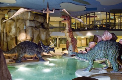 Abendliche Aufnahme aus dem Kinderbereich. Vier lebensgroße Dinosaurier stehen drum herum oder fliegen über das Becken.