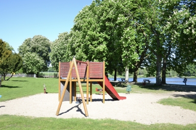 Spielplatz mit Schaukel, Klättergerüst und Rutsche mit Bäumen im Hintergrund.